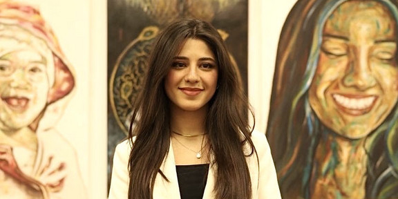 Hales Murad hat lange dunkle Haare und trägt einen hellen Blazer. Sie steht vor einer Wand, an der gemalte Bilder hängen. Sie lächelt.