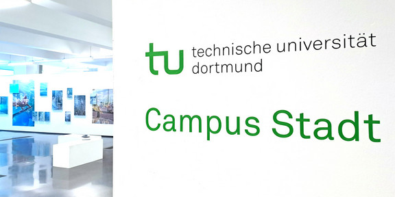 Foto: Rechts im Vordergrund Wand mit Logo der TU Dortmund, darunter grüner Schriftzug "Campus Stadt". Links im Hintergrund Aussstellungsraum mit Sitzmöglichkeiten und verschiedenen Bildern an der Wand.