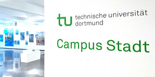 Foto: Rechts im Vordergrund Wand mit Logo der TU Dortmund, darunter grüner Schriftzug "Campus Stadt". Links im Hintergrund Aussstellungsraum mit Sitzmöglichkeiten und verschiedenen Bildern an der Wand.