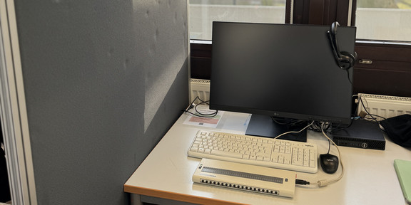 Foto von einem Tisch vor einer Fensterreihe. Auf dem Tisch Bildschirm, PC, Tastatur, Headset und Braillezeile. Links eine Trennwand.