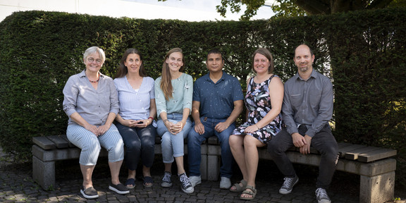 Die 6 Mitarbeiter*innen des Kompetenzzentrums sitzen nebeneinander auf einer Bank und schauen frontal in die Kamera, im Hintergrund: eine Hecke und ein kleiner Ausschnitt des Uni-Gebäudes.