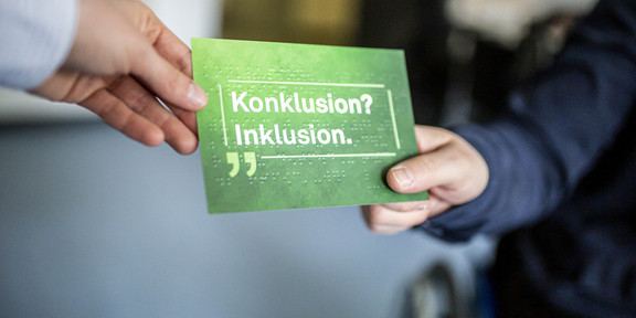 Eine grüne Postkarte mit der Aufschrift „Konklusion? Inklusion.“. Der Schriftzug ist zusätzlich in Brailleschrift in die Postkarte geprägt. An der oberen linken Ecke der Postkarte eine Hand, die die Postkarte an eine Person übergibt, die die rechte Ecke hält. Im Hintergrund das Rad eines Rollstuhls.