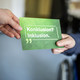 Eine grüne Postkarte mit der Aufschrift „Konklusion? Inklusion.“. Der Schriftzug ist zusätzlich in Brailleschrift in die Postkarte geprägt. An der oberen linken Ecke der Postkarte eine Hand, die die Postkarte an eine Person übergibt, die die rechte Ecke hält. Im Hintergrund das Rad eines Rollstuhls.