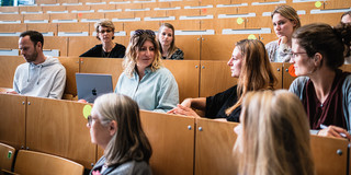 Zwei weiblich gelesene Personen reden mit einander in einem Hörsaal. Andere Kommiliton*innen hören den beiden zu und schauen sie an.