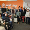 Foto: Eine Gruppe von 18 Personen steht und sitzt in einem Büro vor einer orangefarbenen Wand mit dem weißen Logo und Schriftzug "rapidminer". Die Personen lächeln in die Kamera und tragen überwiegend legere Kleidung. Im Vordergrund sitzen zwei Personen auf einem weißen Sofa, während die restlichen Personen im Hintergrund stehen. Der Raum ist modern eingerichtet und hell beleuchtet.