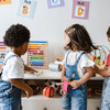 Three toddlers are standing in front of a low shelf with toys. One of the children reaches for a pencil lying on the shelf.