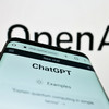 Ein Smartphone, auf dem die Webseite "ChatGPT" geöffnet ist, vor einem Logo von OpenAI.