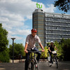 A man with a red helmet is sitting on a bicycle and stops, next to him a woman with a white helmet is riding a bicycle.