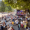 Vor einer Musikbühne sind viele Menschen stehend und auf Bierzeltbänken sitzend versammelt.