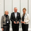 A man wearing a suit stands in the center and holds an award in his hands. To his left stands an elderly lady wearing many pearl necklaces and formal clothing. To his right, a woman wearing a white blazer and a black dress.