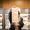 A young woman stands in front of a canteen shelf filled with sandwiches and holds a sandwich bag with the words "Violence doesn't come on campus" in front of her face.