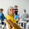 Eine Frau mit grauen Haaren sitzt in einem Seminarraum und lächelt über die Schulter in die Kamera. Im Hintergrund sitzen weitere Teilnehmende, die einer stehenden Dozentin zuhören. Vor der Frau steht ein Laptop.