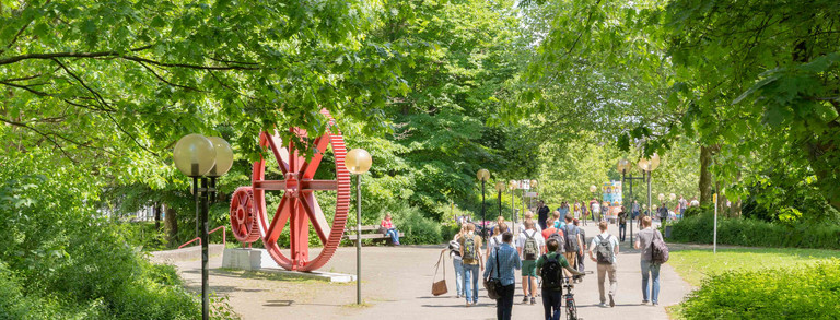 Studierende sind im Sommer auf dem Campus Nord, rote Zahnräder im Hintergrund.