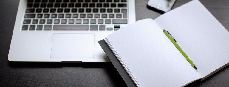 Laptop, Smartphone und Notizbuch mit Stift auf einem Tisch liegend.