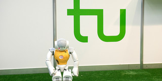 Ein Roboter mit gelbem Trikot steht an der Seitenlinie eines Fußballfelds, hinter ihm ist das grüne Logo der TU Dortmund.