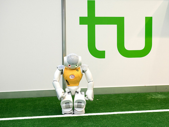 Fußballroboter vor TU-Logo Ein Roboter mit gelbem Trikot steht an der Seitenlinie eines Fußballfelds, hinter ihm ist das grüne Logo der TU Dortmund.
