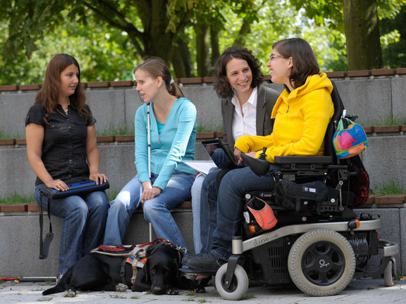 Vier Studierende, darunter Studierende mit Behinderung, sitzen gemeinsam auf dem Campus.