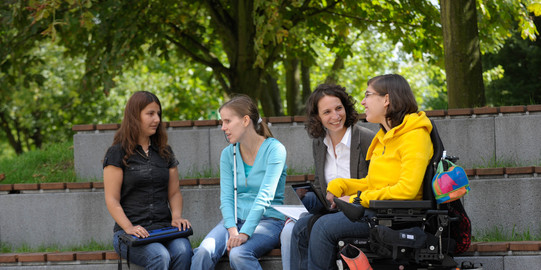 Studierende mit Behinderung Vier Studierende, darunter Studierende mit Behinderung, sitzen gemeinsam auf dem Campus.