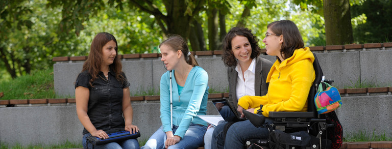 Studierende mit Behinderung Vier Studierende, darunter Studierende mit Behinderung, sitzen gemeinsam auf dem Campus.