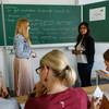 Two persons are standing in front of a board with formulas on it. In the foreground there are three students working at desks.