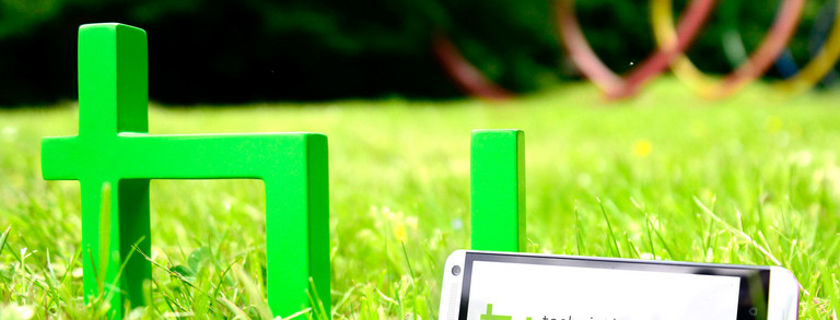 The green logo of TU Dortmund University is standing on a meadow. A smartphone is leaning against it.