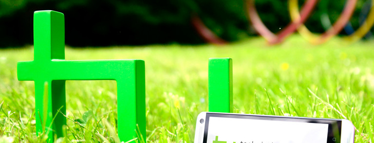 Ein grünes TU-Logo steht auf grünem Rasen. Daran lehnt ein Smartphone.