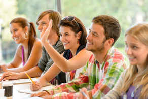 Two boys and two girls sit next to each other at a table in the classroom and raise their hands.