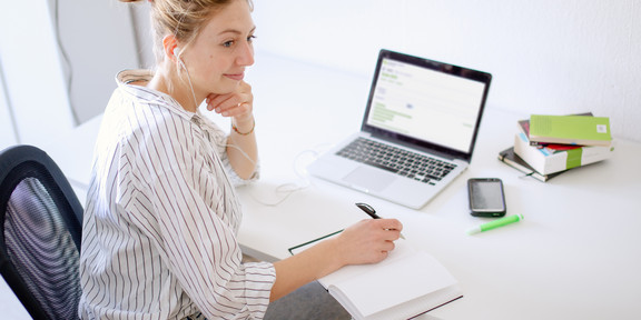 Woman sitting at her desk with a laptop writing something down in a notebook